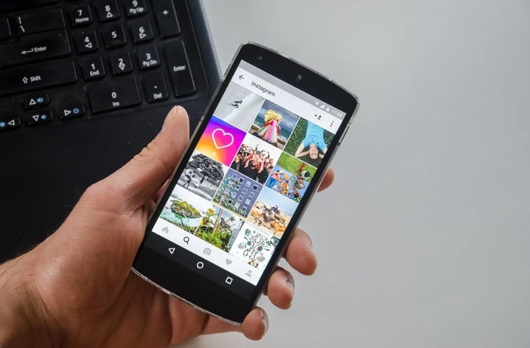 На фоне ноутбука человек держит в руке смартфон с запущенным приложением Instagram